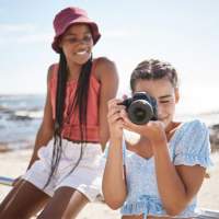 Two young women on a beach, one smiling while the other takes a photo with a camera. The ocean is visible in the background.