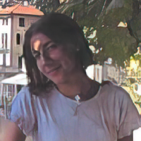 A woman in a white shirt stands outside with buildings and trees in the background.